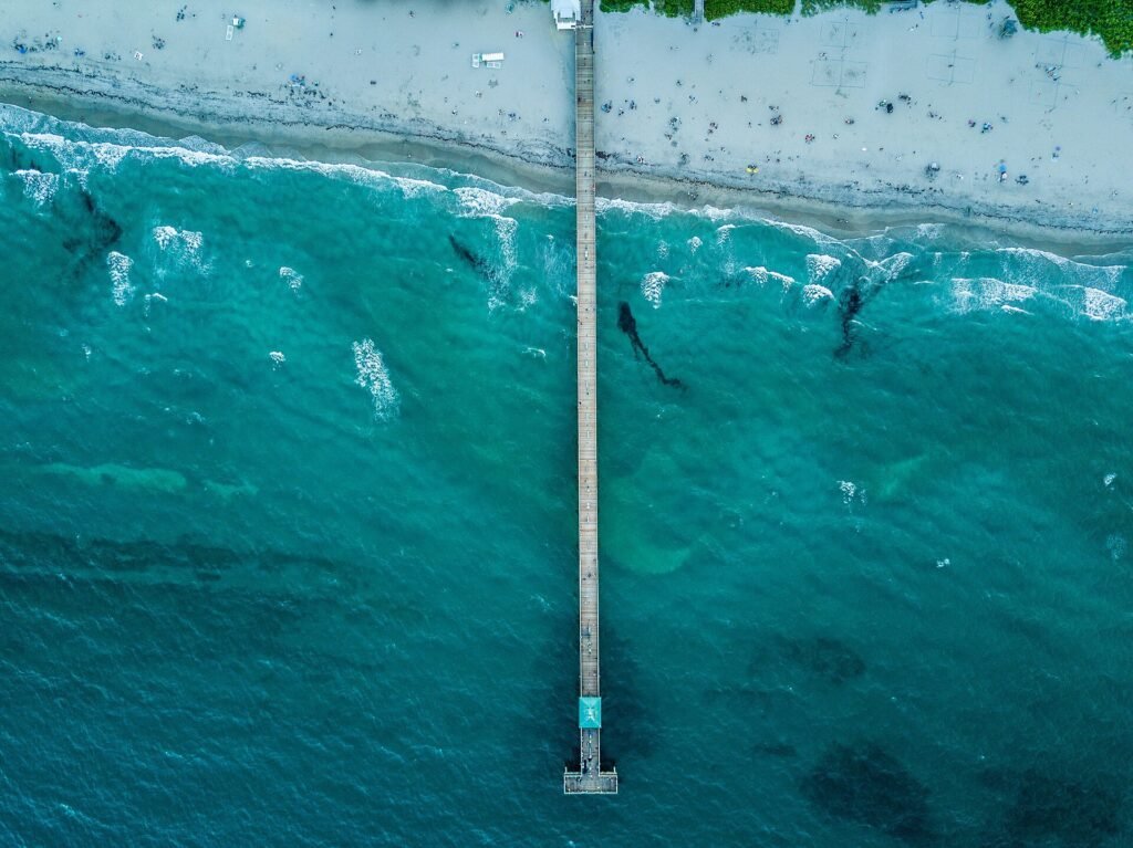 Aerial view of Deerfield Beach Pier extending into the Atlantic Ocean, with the surrounding beachfront and coastal waters.