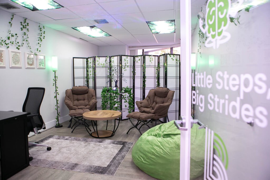 Two cushioned chairs, a round table and a green beanbag in a wellness room, viewed through a Little Steps, Big Strides door.