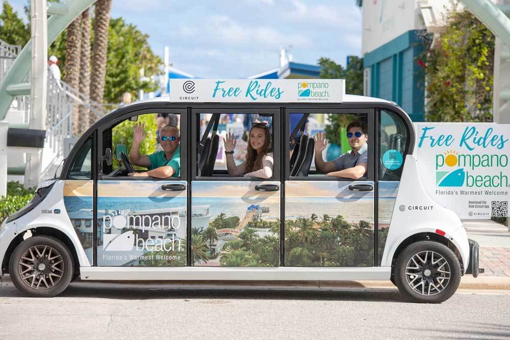 Three people wave from a white Pompano Beach Circuit shuttle displaying Free Rides signs and beach imagery.