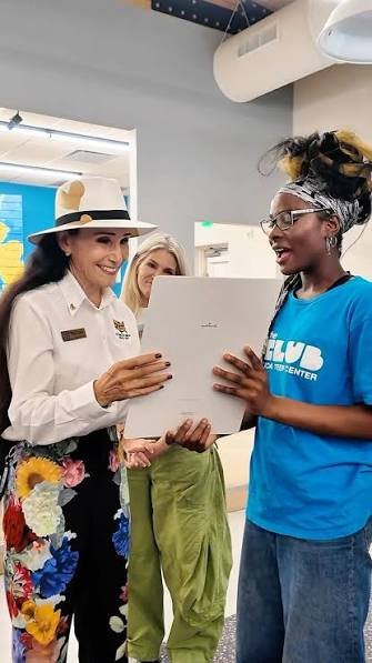 A woman wearing a white hat presents a white package to a young participant in a blue Boys & Girls Clubs shirt while another woman looks on.